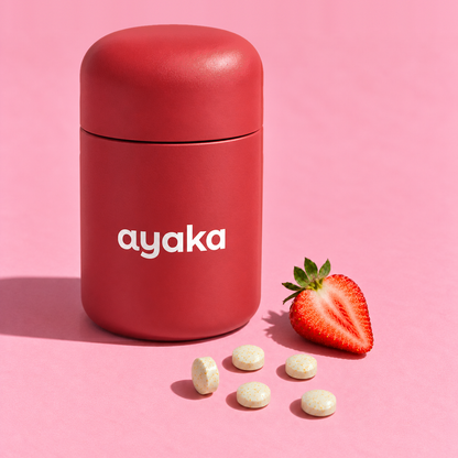 Punainen Ayaka-purkki vaaleanpunaisella taustalla, edessä vitamiinitabletteja ja puolikas mansikka